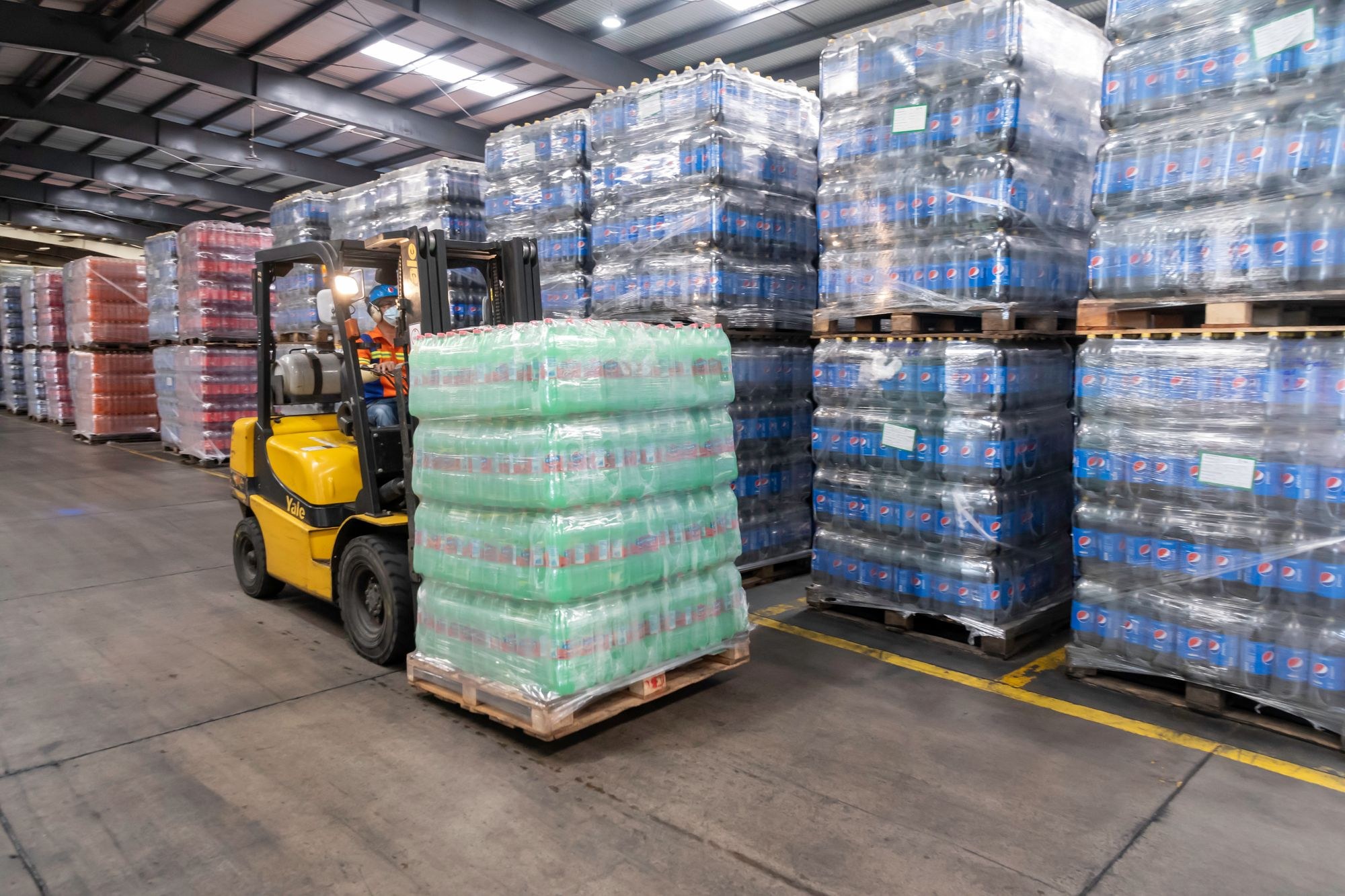 A warehouse forklift operator moving a wrapped pallet of bottled drinks beside rows of stored beverage pallets.
