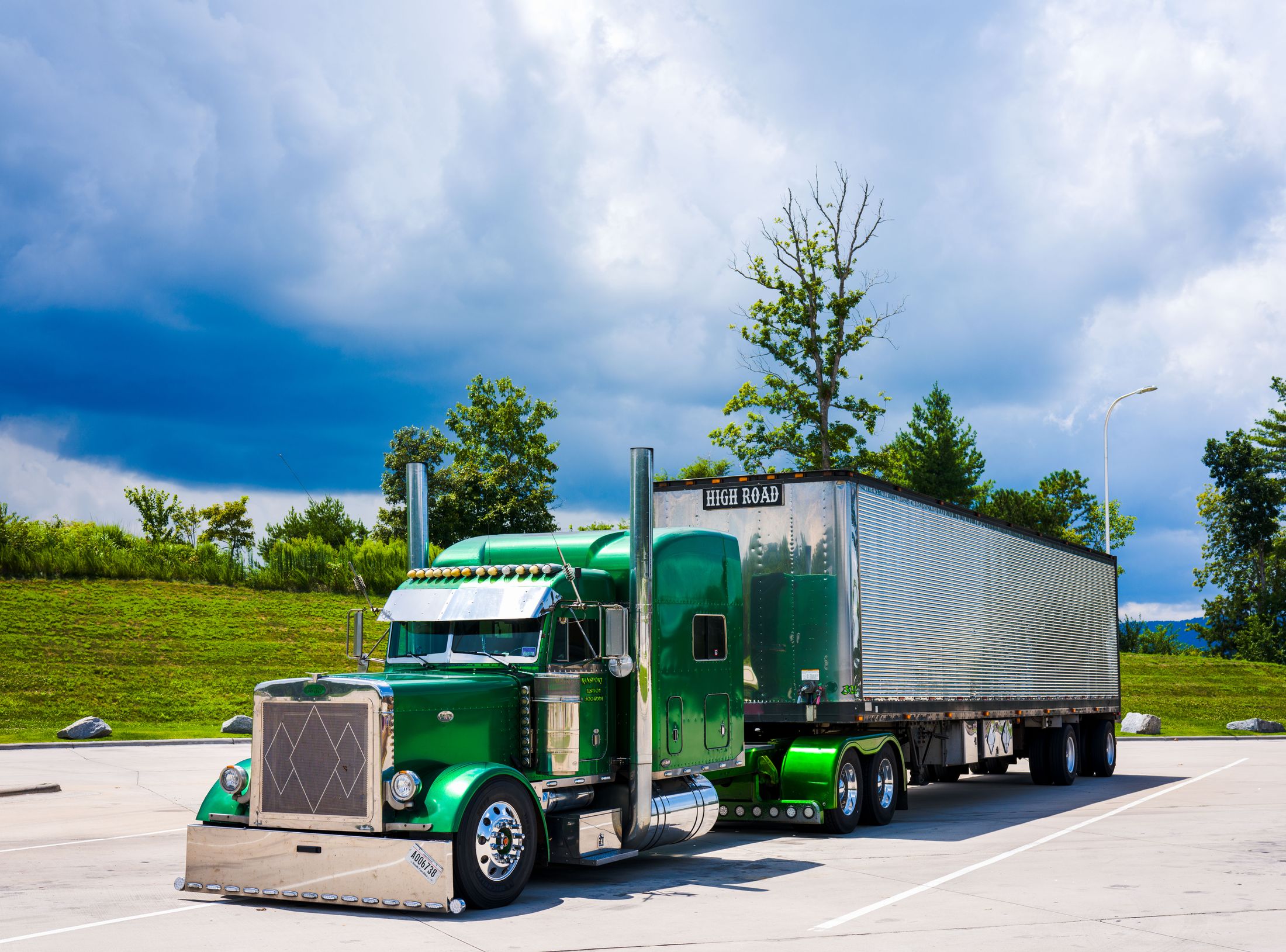 A green American semi-truck with a silver trailer parked beside a grassy, tree-lined embankment