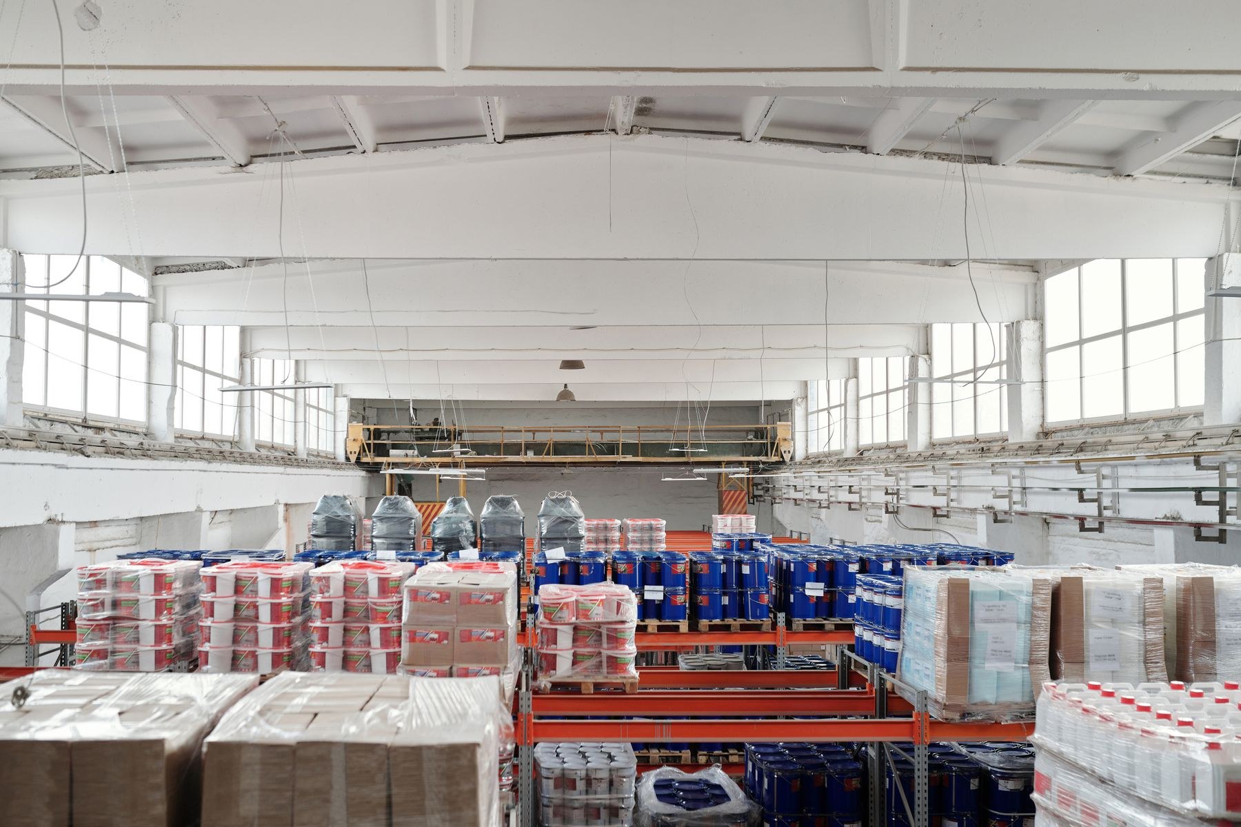 Bright warehouse interior with shrink-wrapped pallets, cartons and blue drums on storage racks.