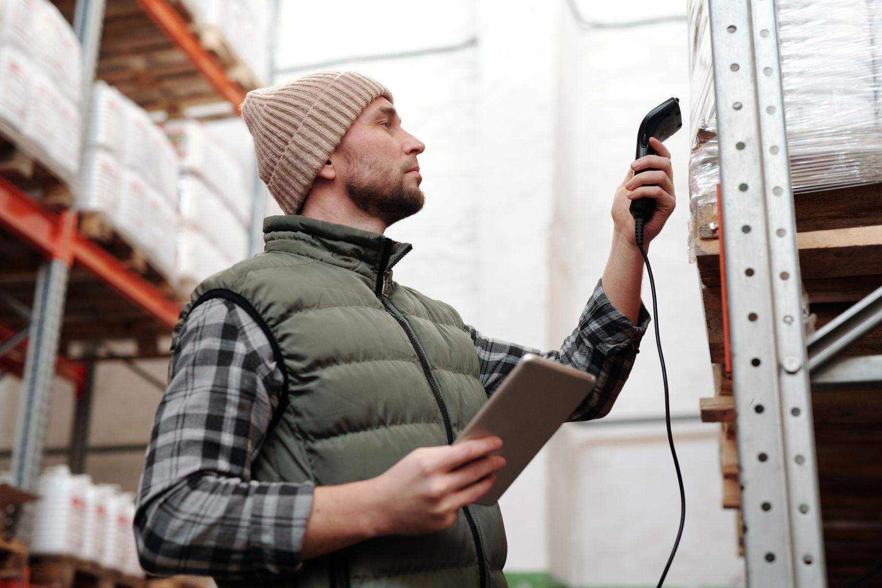 A warehouse worker in a green vest scanning stored inventory while holding a tablet.